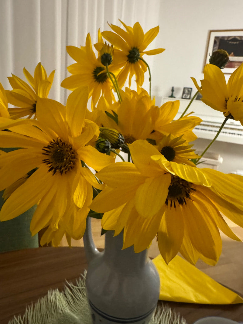 Ein bunter Strauß leuchtend gelber Topinambur-Blüten steht dekorativ in einer hellen Keramikvase auf einem Tisch im Innenbereich.
